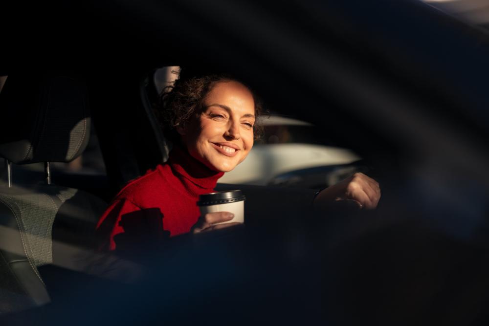 Salary sacrifice as a commuting cost solution: A smiling woman in a red roll-neck jumper holding a takeaway coffee cup whilst sitting in the driver's seat of a car, photographed through the window in warm sunlight.