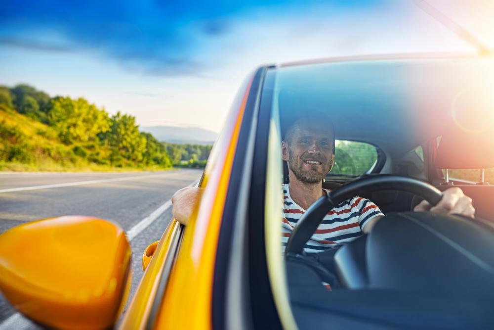 Short-term vehicle hire Short-term vehicle hire: A smiling man with a beard driving a yellow car along a country road on a sunny day, viewed through the windscreen with green hills in the background.
